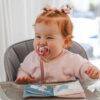Back To Nature Silicone Pacifier Clip in pink, attached to a baby’s pacifier, with a smiling child in a high chair.