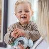 Back To Nature Teether & Spinning Rattle in blue and pink, held by a smiling toddler in a cozy indoor setting.