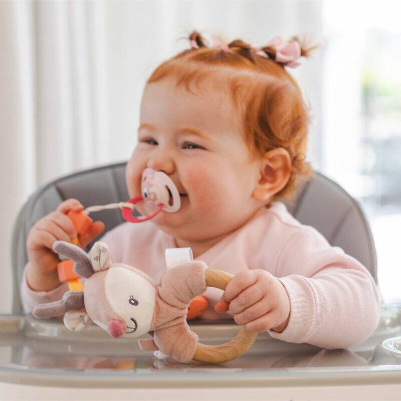Back To Nature Silicone Pacifier Clip in blue and pink, attached to a smiling baby in a high chair, holding a toy.