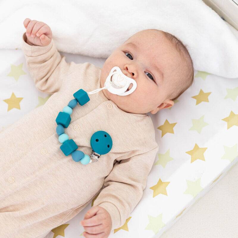Back To Nature Silicone Pacifier Clip in blue and pink, featuring a cube design, attached to a baby’s pacifier.