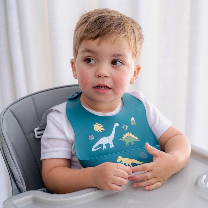 Blue Back To Nature Silicone Bib featuring dinosaur designs, worn by a child sitting in a high chair.