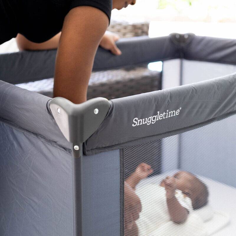 Snuggletime Classic Comfort Camp Cot in gray, with a parent gently placing a baby inside the cot's mesh sides.
