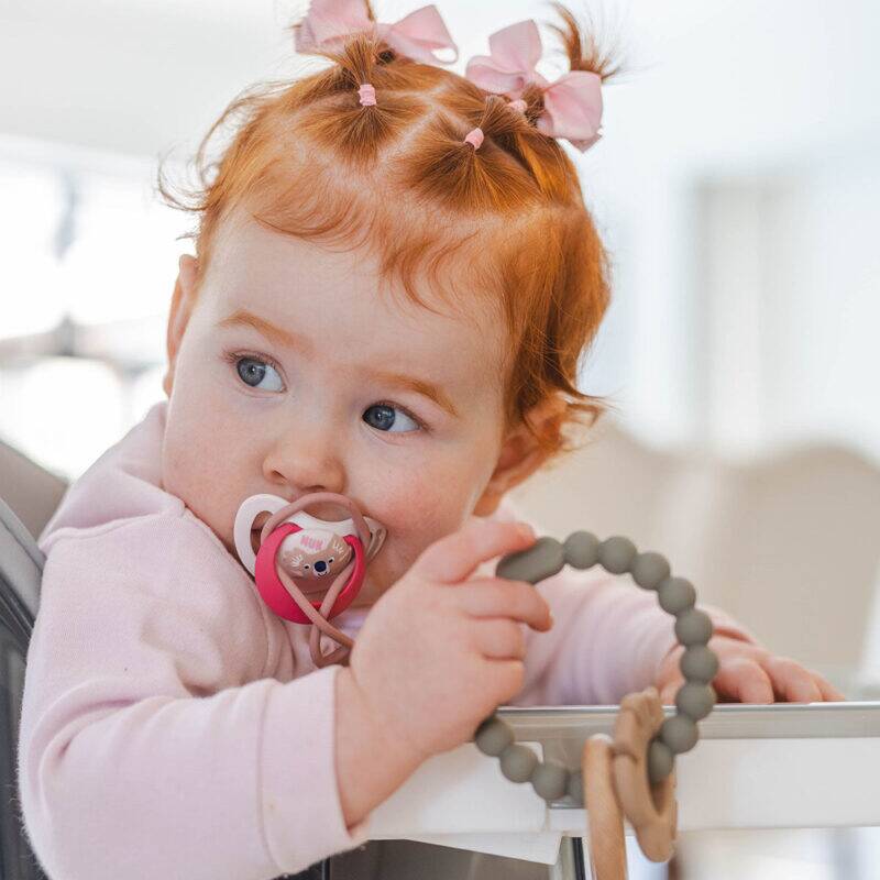 Baby girl in a high chair holding a Back to Nature Silicone Teether Leaf in a soft green color, with a pacifier in her mouth.