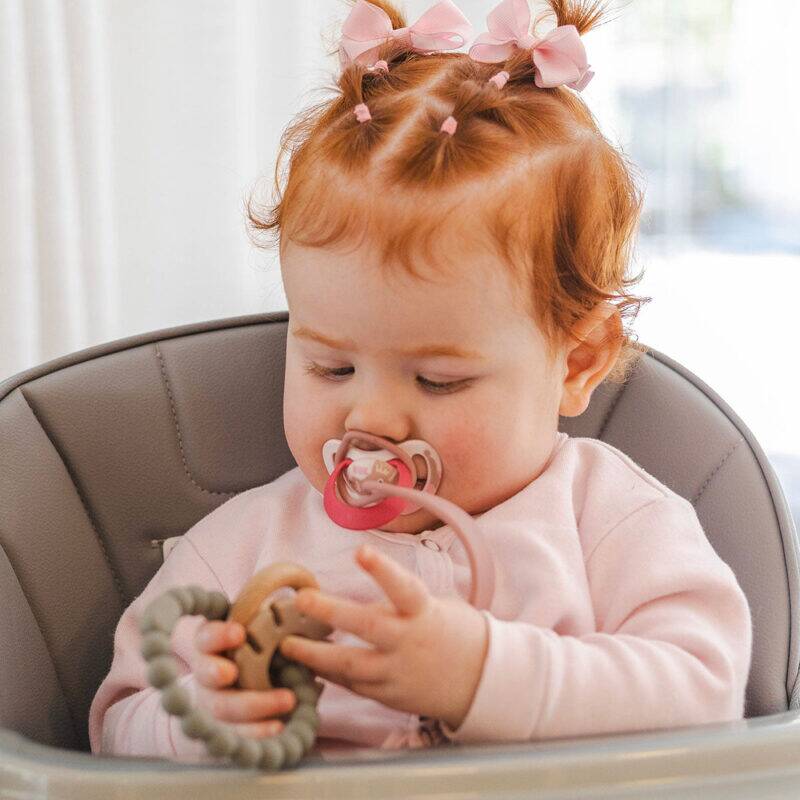 Back to Nature Silicone Teether Leaf in use by a baby with red hair in a high chair, exploring the teether with curiosity.