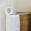 BabySafe Nappy Bin Deodoriser in white, placed on a floor next to a woven basket, with lid open showing deodorising feature.