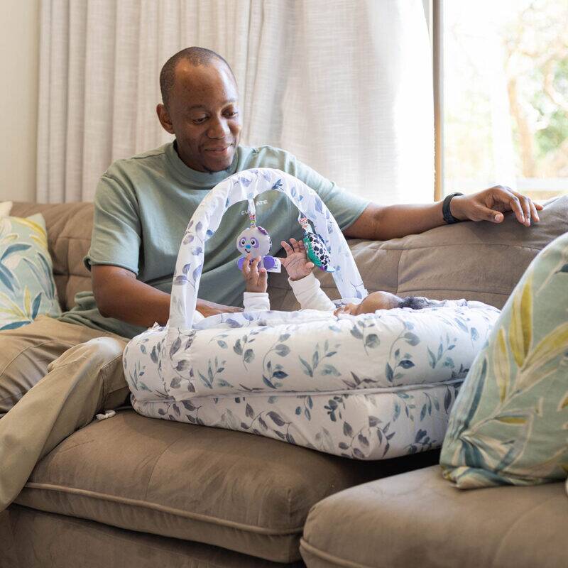 Snuggletime Deluxe Comfort Pod with Activity Arch, featuring a parent playing with a baby, set on a cozy couch.