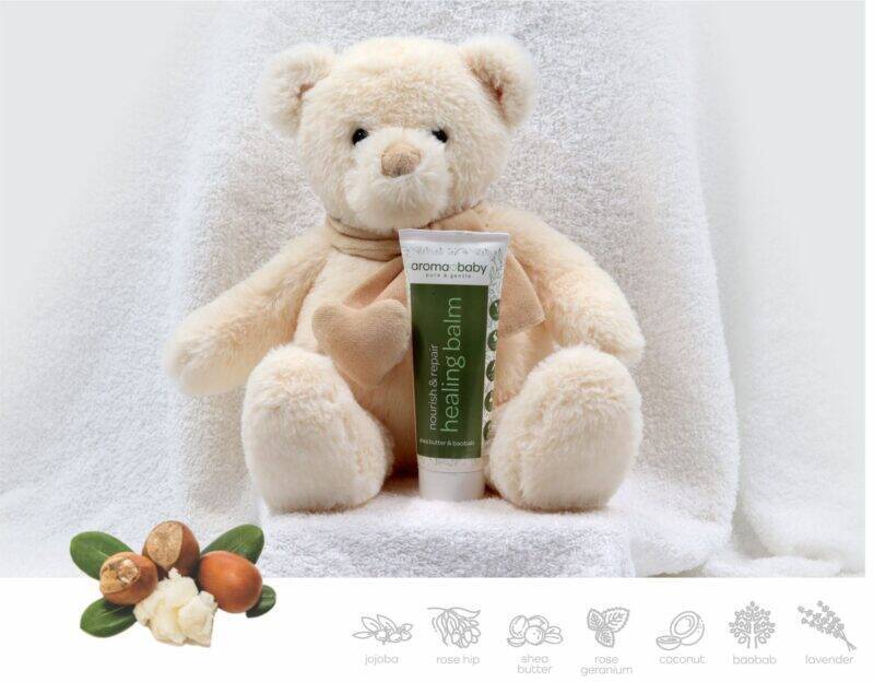 Aromababy Healing Balm tube displayed with a soft teddy bear against a white towel backdrop, highlighting its soothing