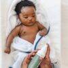 Aromababy baby lotion held by an adult hand, with a baby wrapped in a white towel on a soft surface.