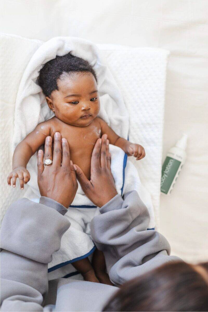 Aromababy baby lotion being applied to a baby's chest by a caregiver, with a towel and lotion bottle nearby.