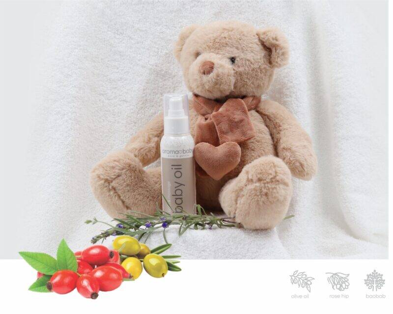 Aromababy baby oil bottle next to a soft teddy bear, set against a cozy white backdrop with natural elements.