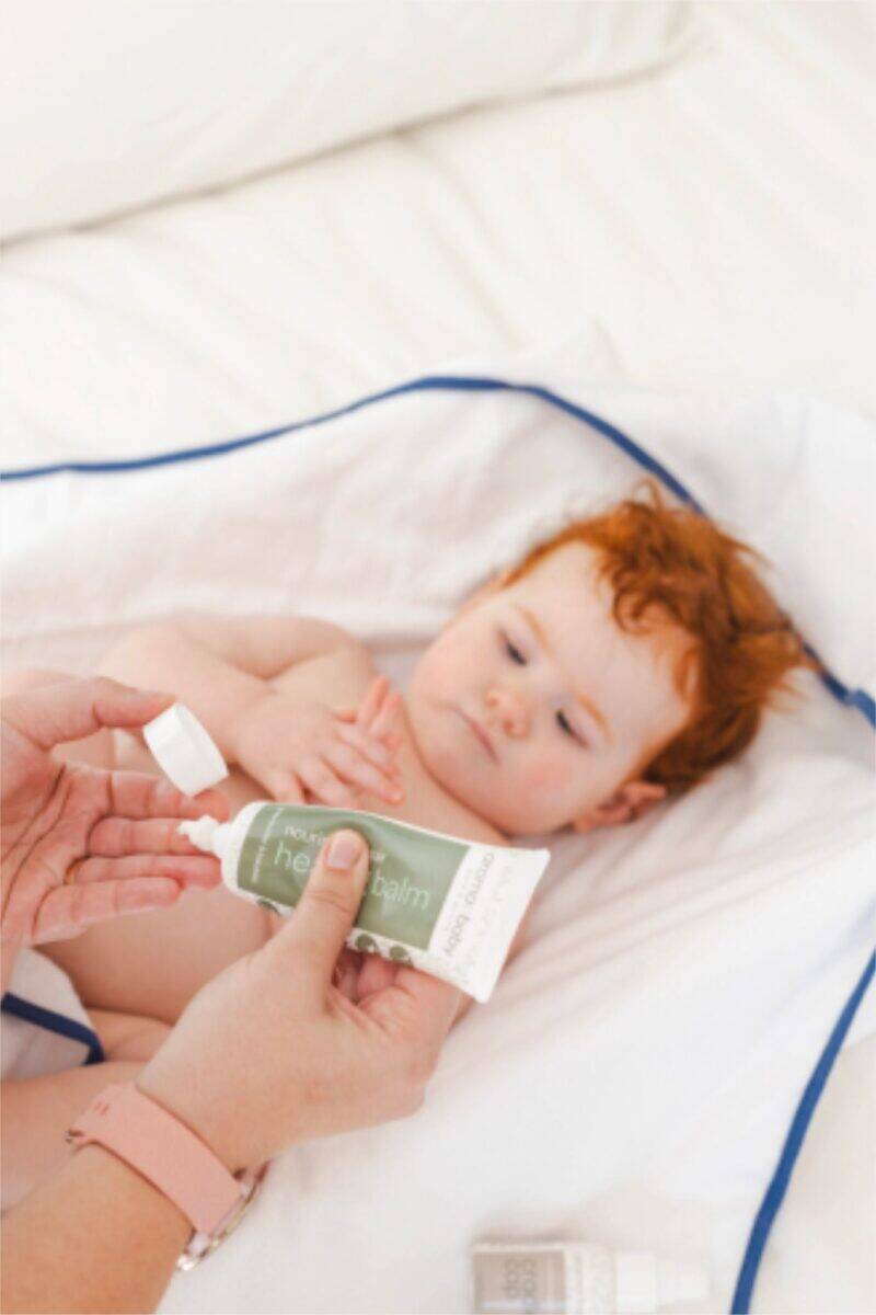 Aromababy Healing Balm tube held by an adult, with a baby lying on a white blanket, looking curiously at the balm.