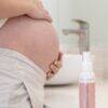 Aromababy mom-to-be essentials stretch mark spray next to a pregnant woman's belly, set in a bright bathroom.