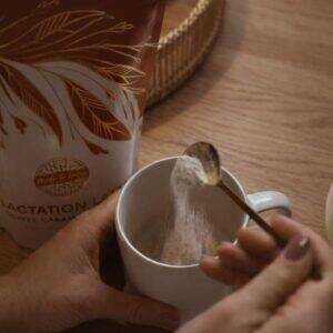 Proud to Love Lactation Latte in Toffee Caramel flavor, being stirred into a white mug with a wooden spoon.
