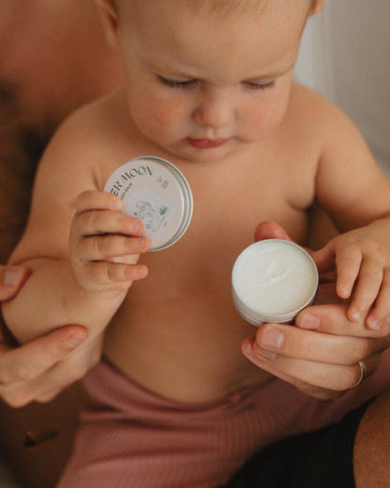 Baby holding a small cream container with a floral lid, seated on an adult's lap, wearing pink shorts.