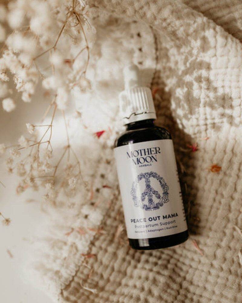 Mother Moon Peace Out Mama postpartum support tincture in a dark bottle, placed on a textured beige fabric with dried