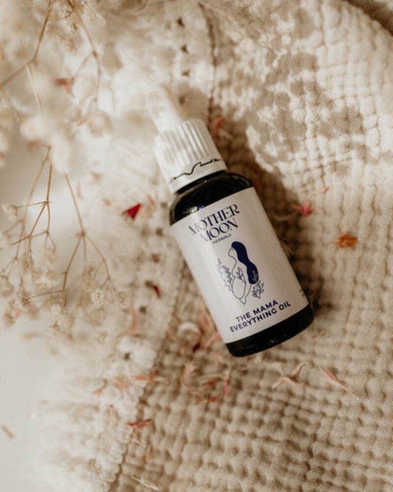 Mother Moon The Mama Everything Oil in a dark glass bottle with a white cap, placed on a textured cream fabric.