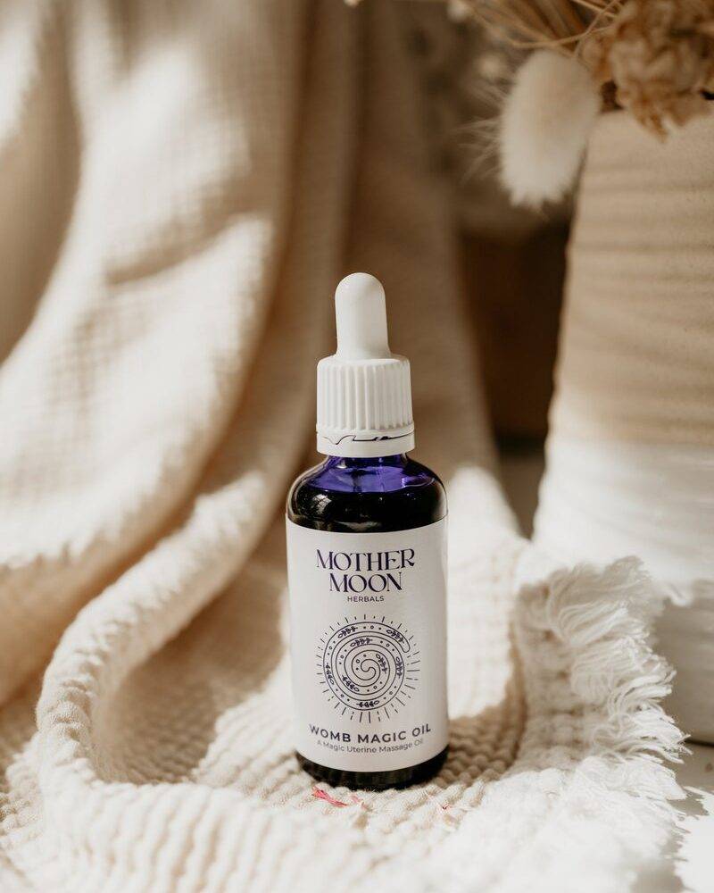 Mother Moon Womb Magic Oil in a blue glass dropper bottle, placed on a textured beige blanket with dried flowers in the