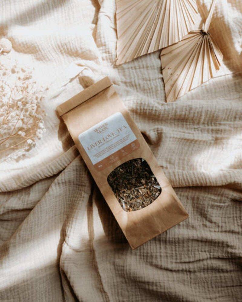 Lorna Jane herbal tea blend in a kraft paper bag with a clear window, placed on a textured beige fabric backdrop.