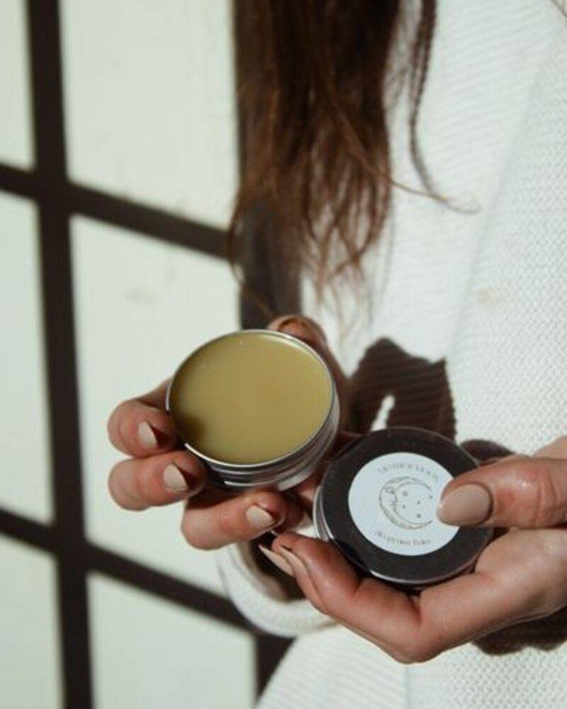Natural skincare balm in a silver tin held by a person, with a light green color and minimalist label design.